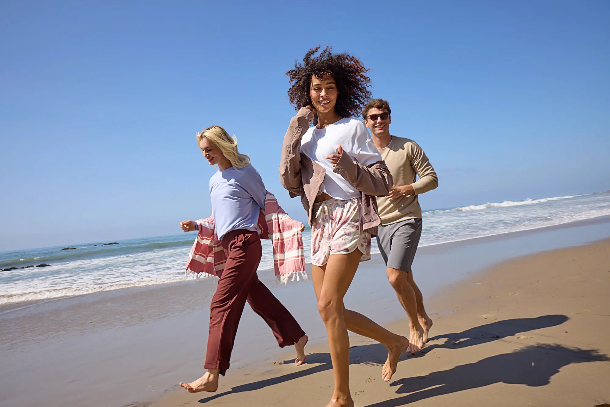Two women and a man, happily walking barefoot on a sandy beach, with blue sky and rolling waves in the background