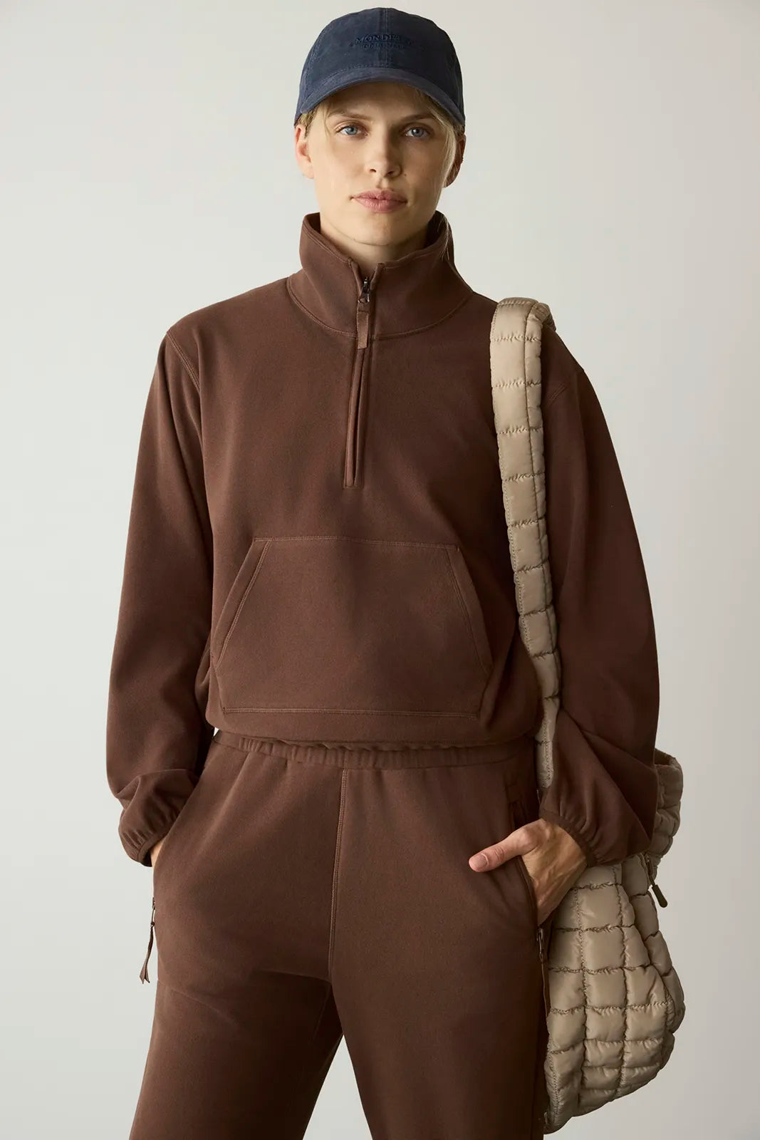 A model wearing a matching brown quarter-zip pullover and sweatpants set, styled with a navy blue beanie and a beige quilted shoulder bag against a light gray studio background.