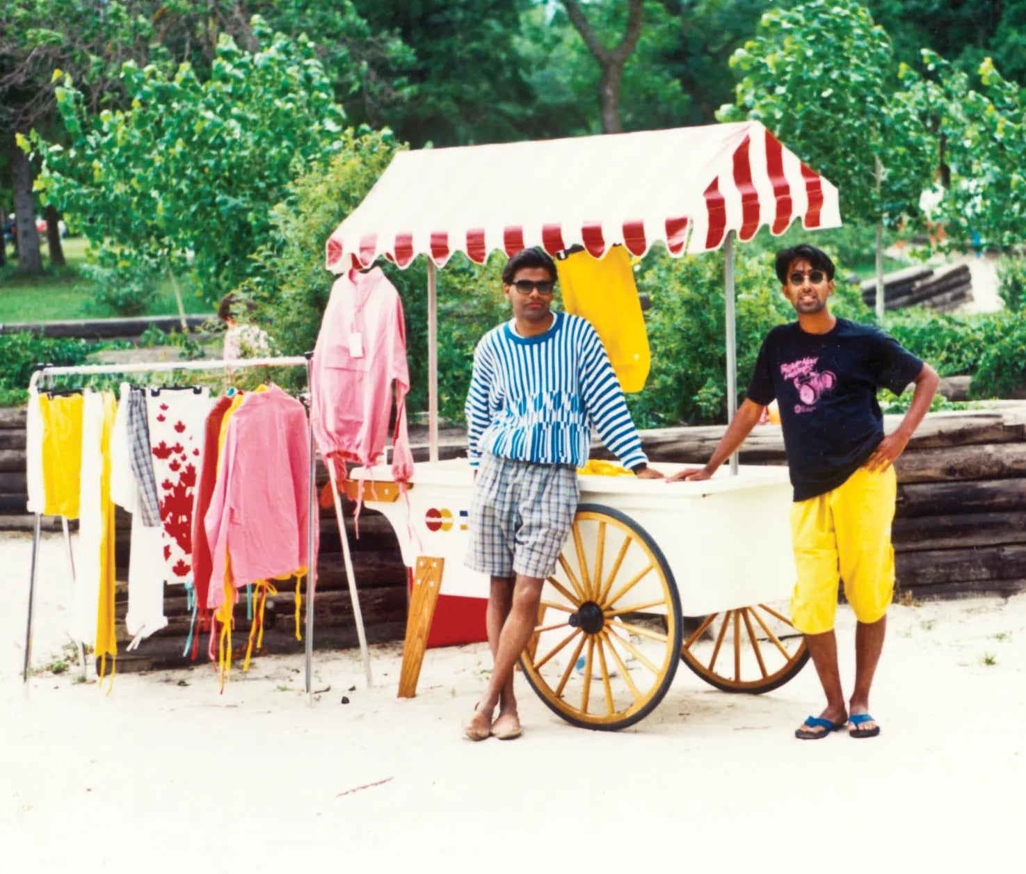 Two Indian men standing next to a cart with early Mondetta clothing on display outdoors at Grand Beach, Manitoba in the late 1980's