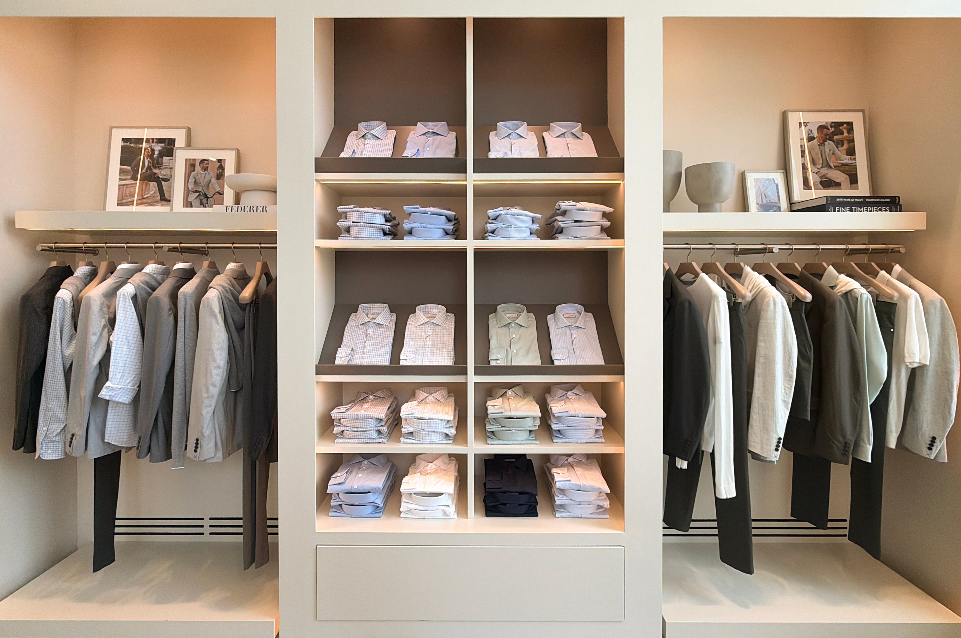 Three thick column cream-color display shelves, center with folded collar shirts, right and left mirrored with vases and photographs on the top shelves and various outfitting hung below.