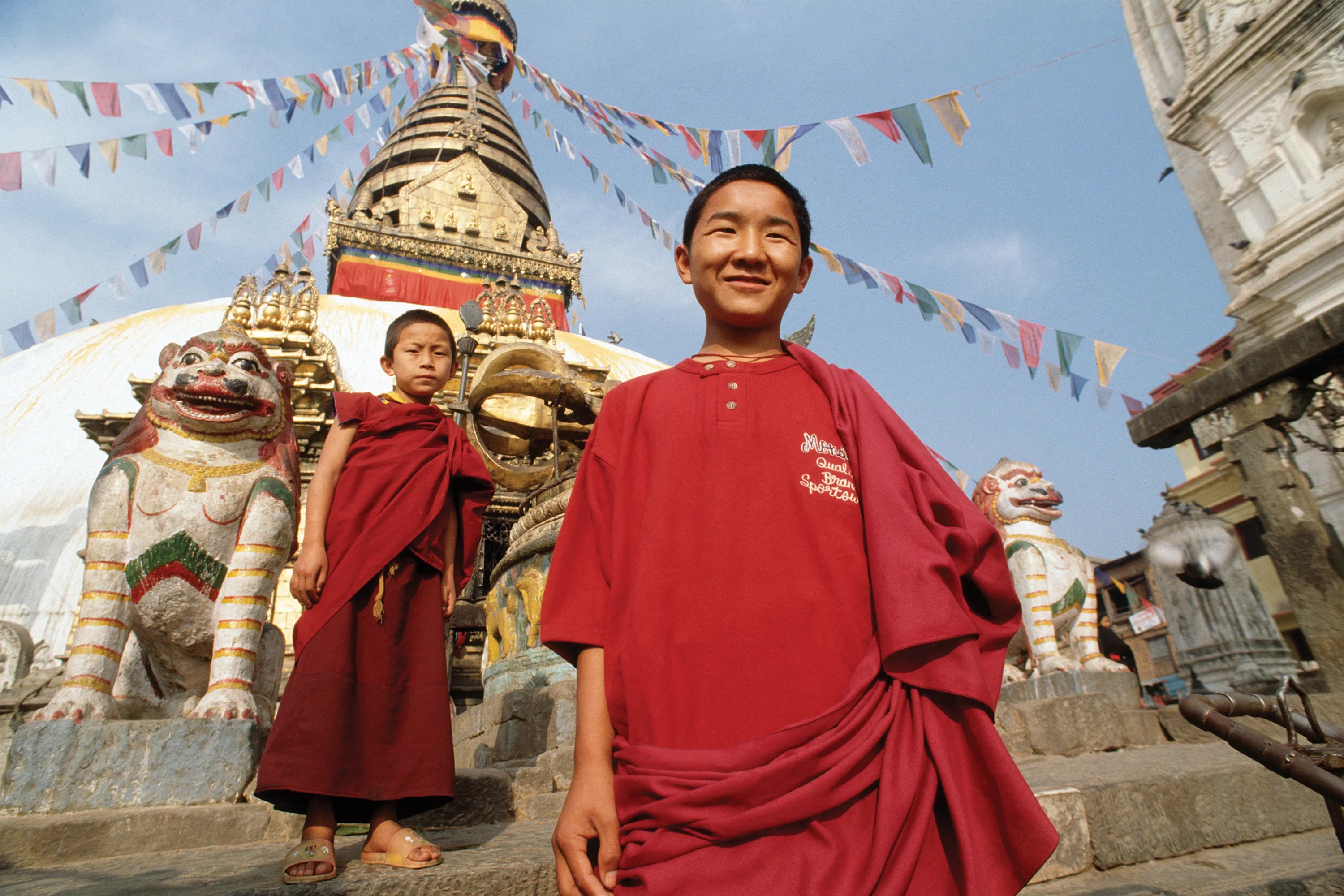 Two young Buddhist boys in red robes and red Mondetta shirts standing in front of a Buddhist temple in China with colorful flags in the background.