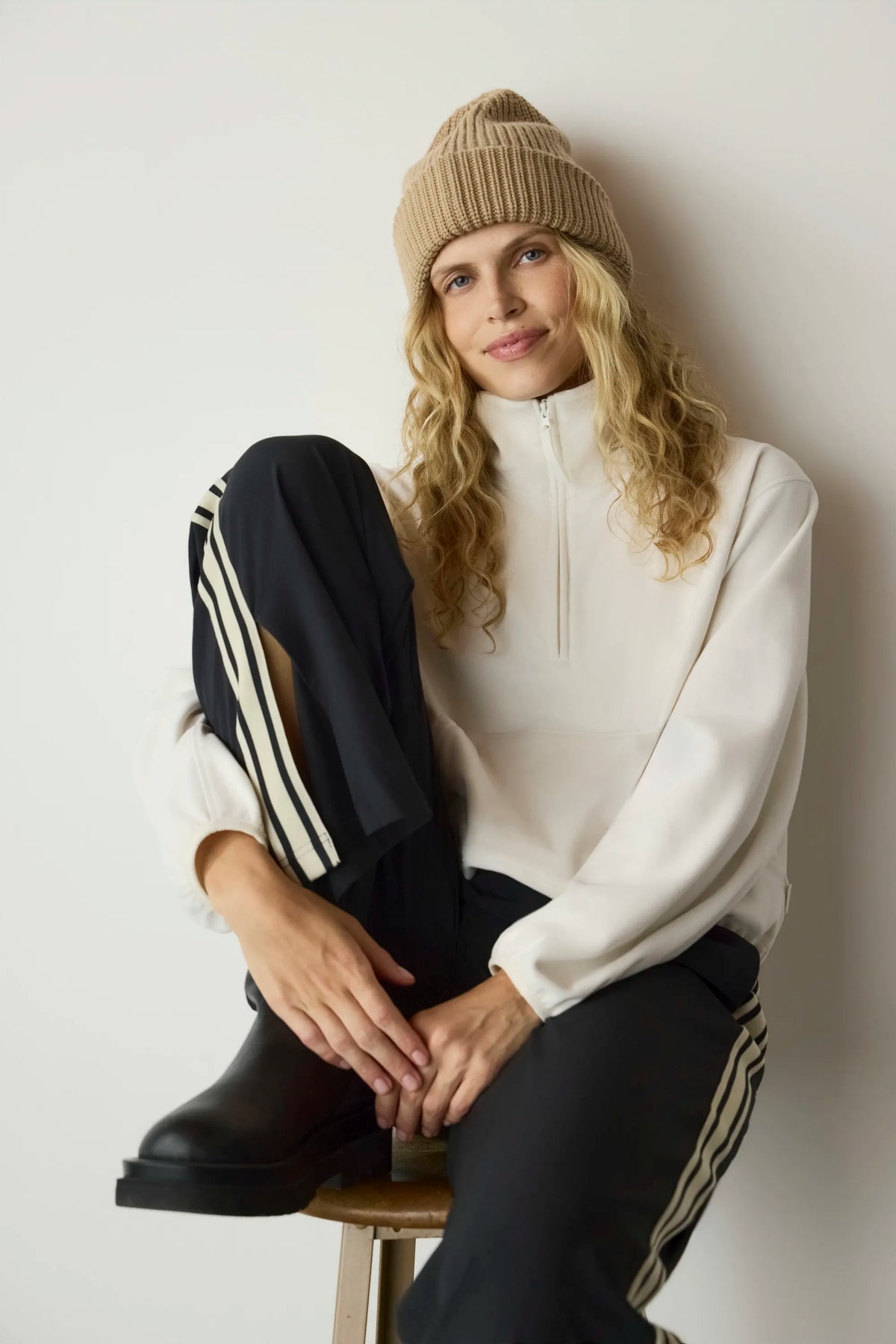 A woman sits on a stool against a plain light wall, wearing a beige knit beanie, a cream half-zip pullover, black track pants with white side stripes, and black boots.