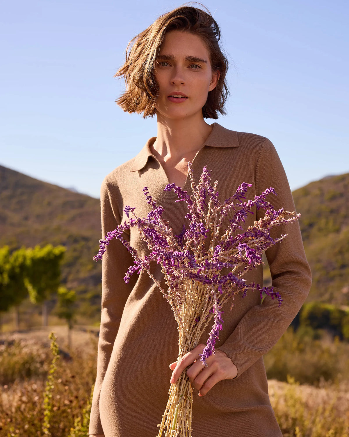 A woman outdoors in a dry grassy landscape wearing a camel brown collared knit sweater dress and holding a bunch of dried purple flowers.
