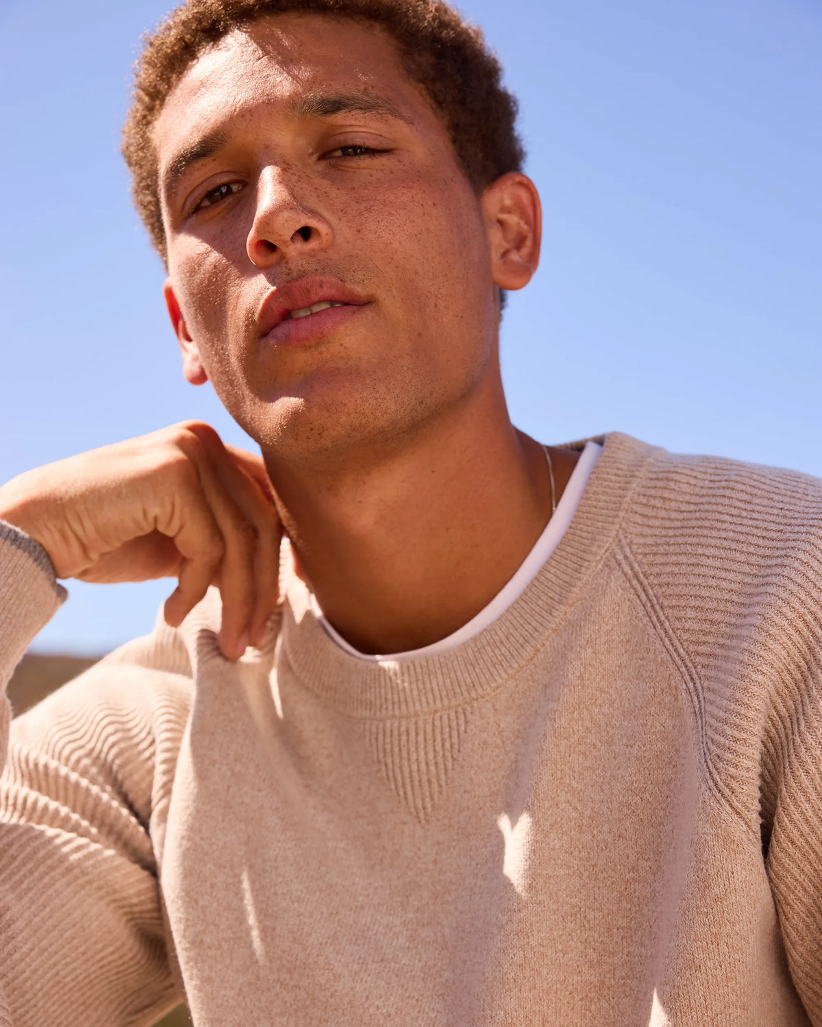A close-up shot of a man wearing a beige ribbed knit crewneck sweater over a white undershirt, set against a bright blue sky background.