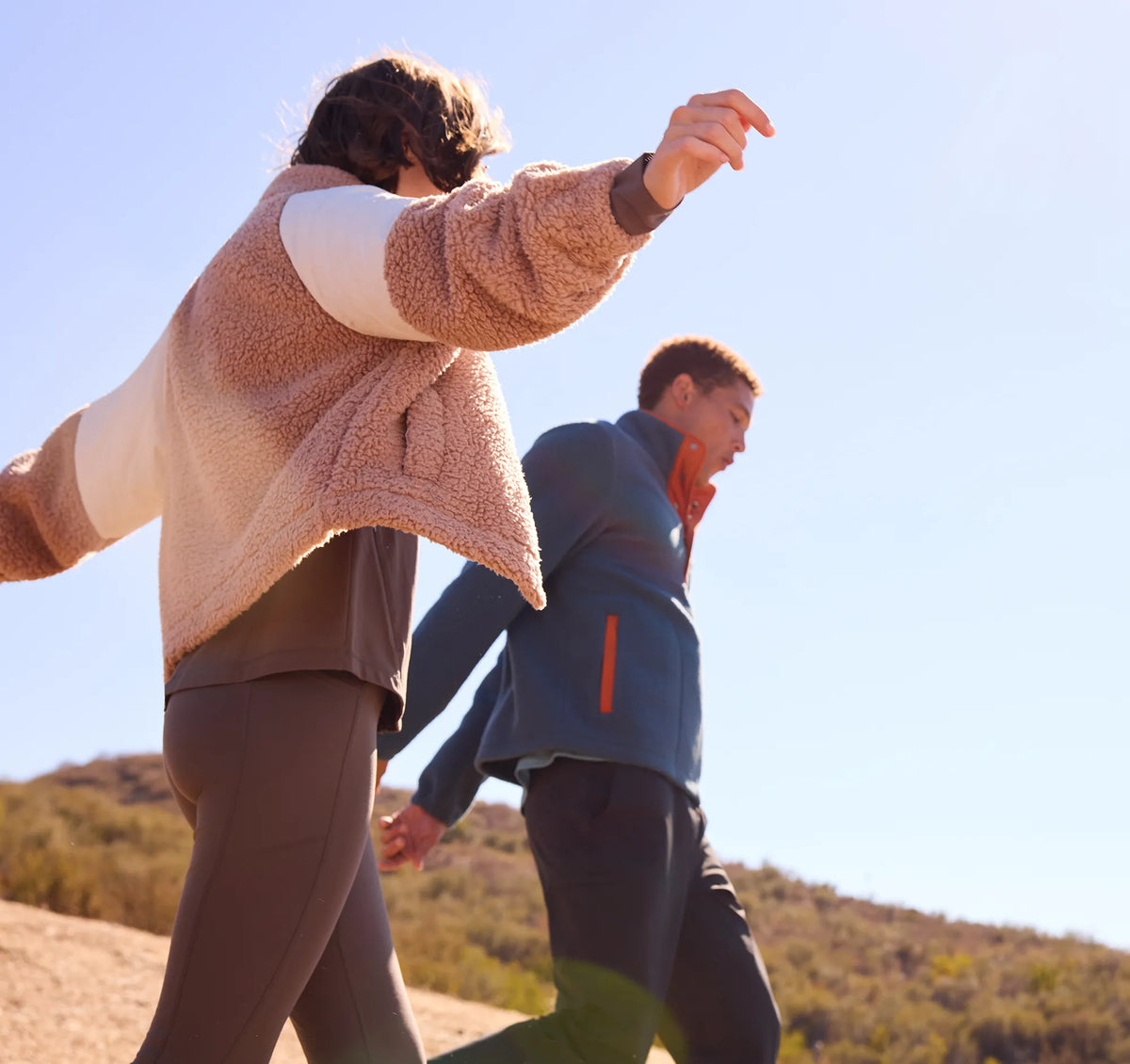 Two people walking outdoors on a sunny day, with the person in front wearing a beige sherpa fleece zip jacket over a white and brown top, and the person behind wearing a blue zip jacket with an orange inner collar.
