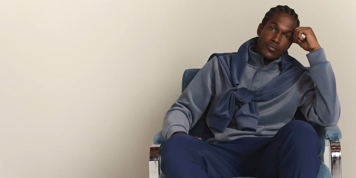A man sits in a blue armchair against a plain beige wall, wearing a matching blue and gray lounge set with a large tied bow at the collar.