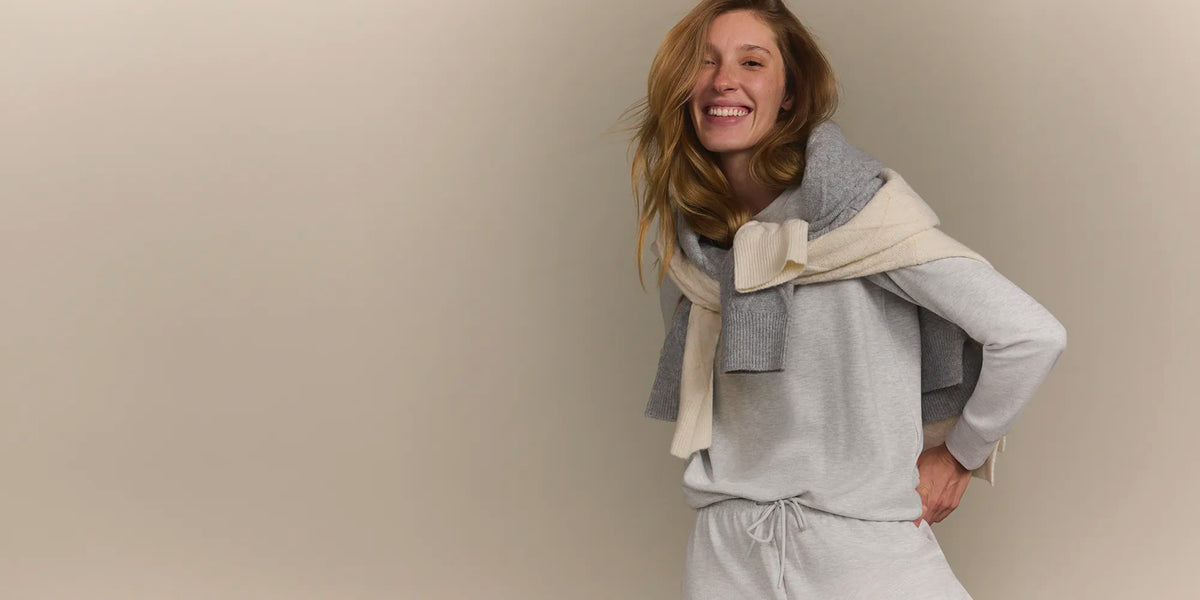 A woman smiling and posing in a light gray sweatshirt and matching sweatpants, with a gray and cream knit scarf wrapped around her shoulders, set against a solid beige studio background.