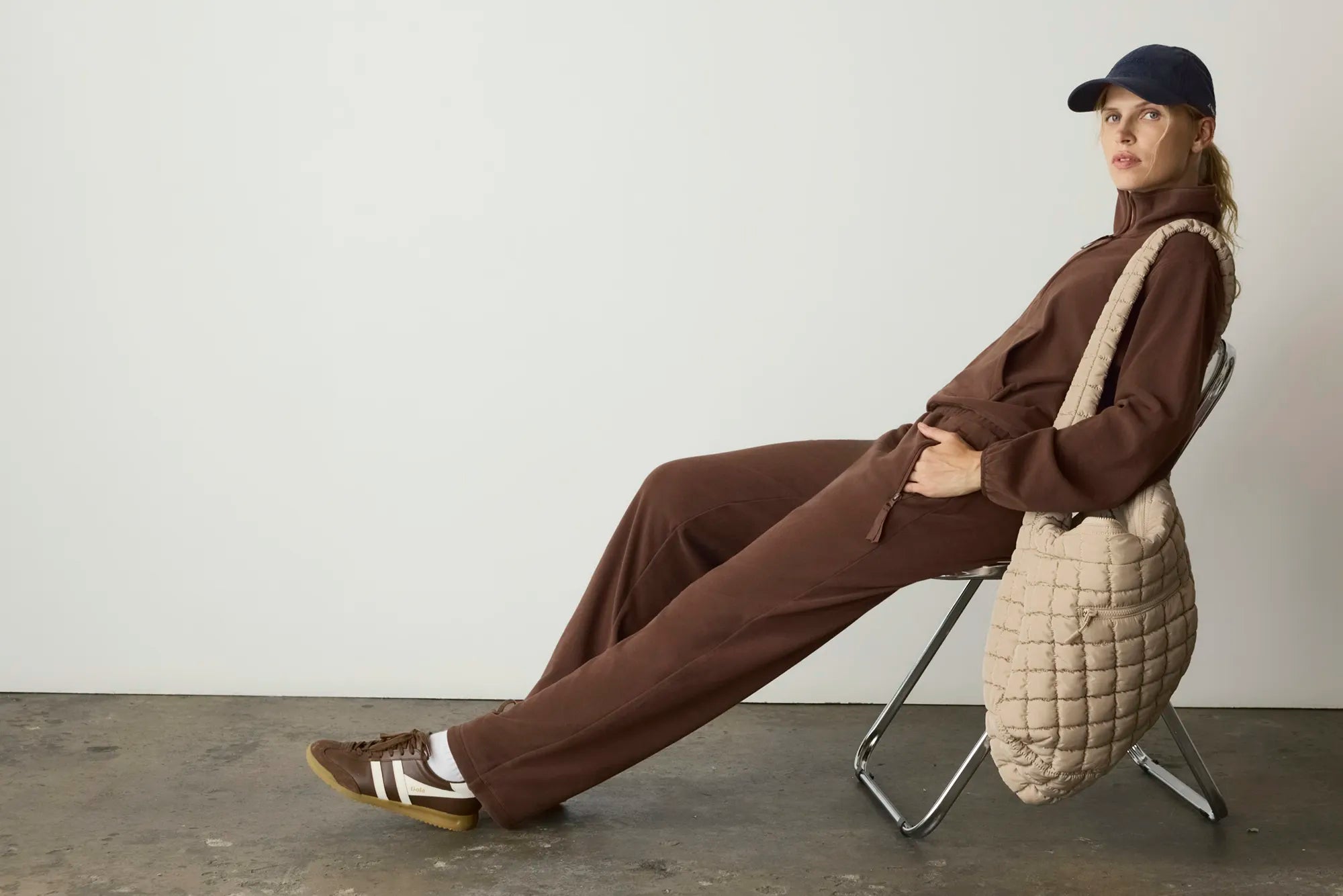 A model sits on a metal folding chair in a minimalist studio setting, wearing a matching brown two-piece tracksuit, brown sneakers, and a navy baseball cap. A large, cream-colored quilted shoulder bag is draped over the chair next to her.