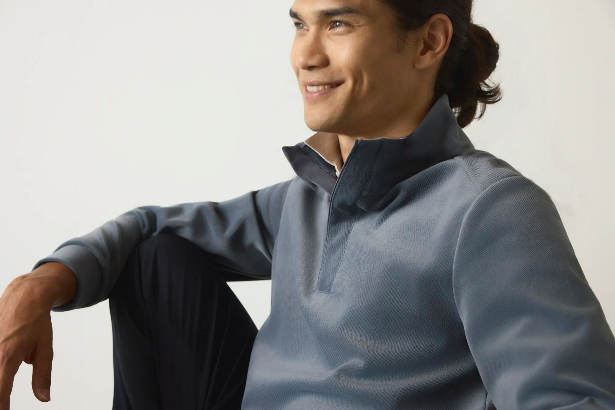 A man wearing a slate blue mock-neck long-sleeve shirt and dark pants, sitting against a plain light background.