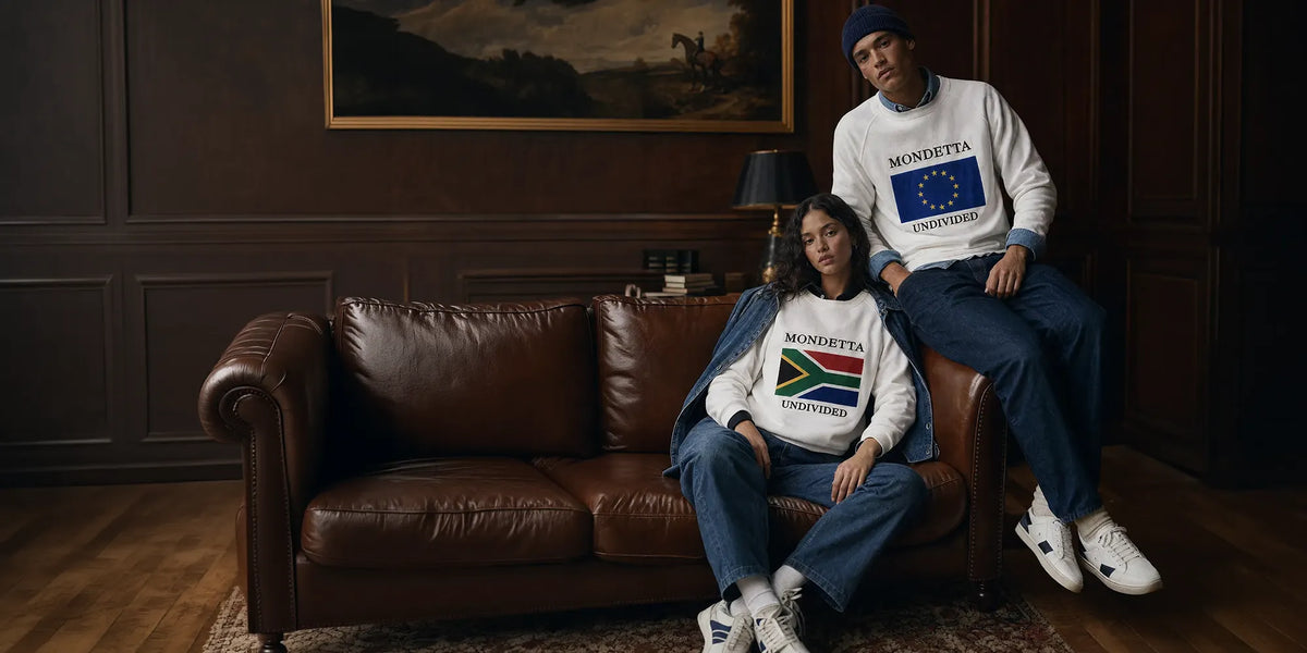 A man and a woman sit on a brown leather couch in a dark wood-paneled room, wearing white crewneck sweatshirts with political slogans. The man's sweatshirt reads 'MONDETTA' with a blue EU flag graphic, and the woman's sweatshirt reads 'MONDETTA' with a South African flag graphic.