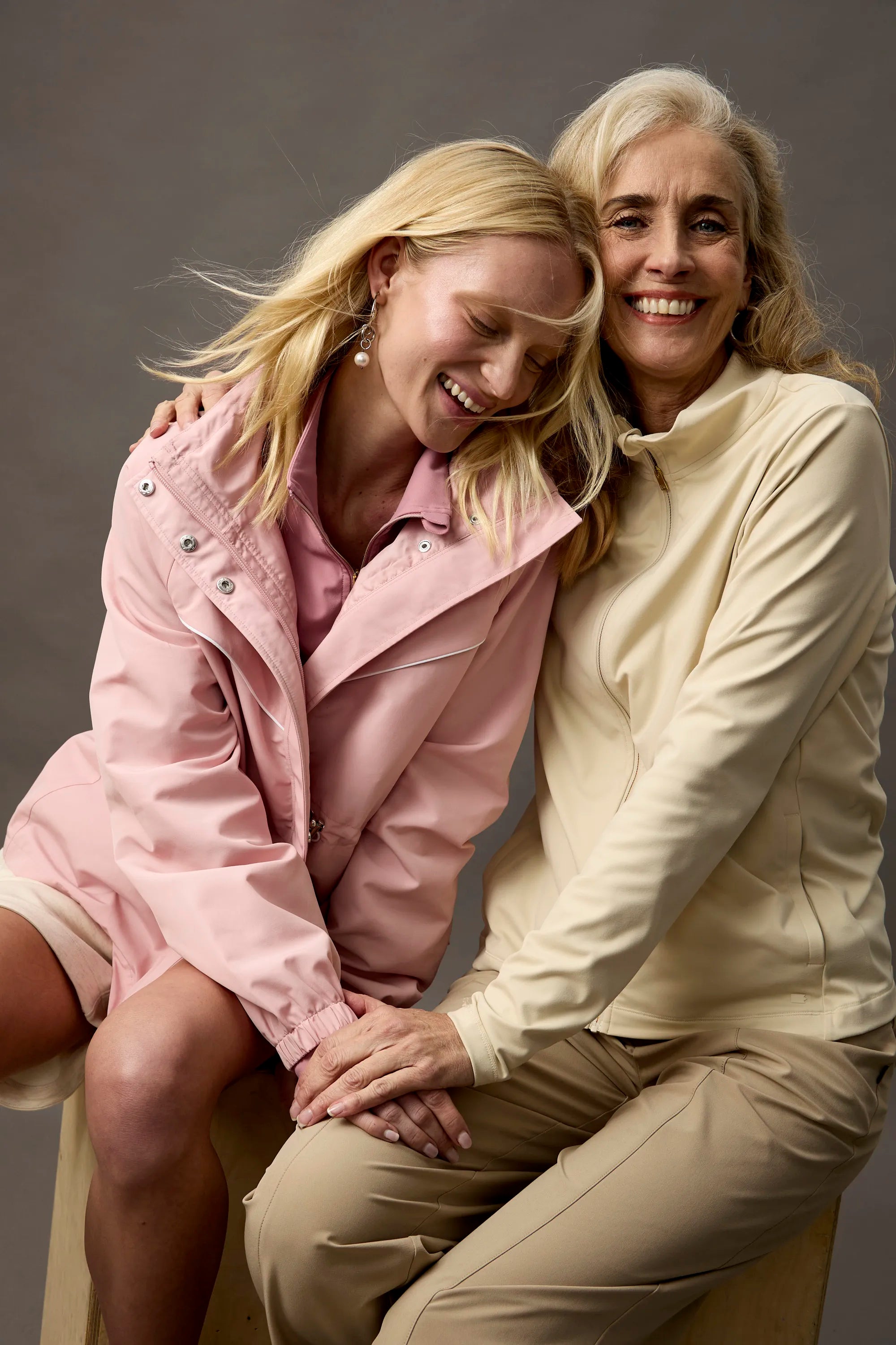 Two women sitting together, one in a pink coat and the other in a beige outfit, against a plain background.