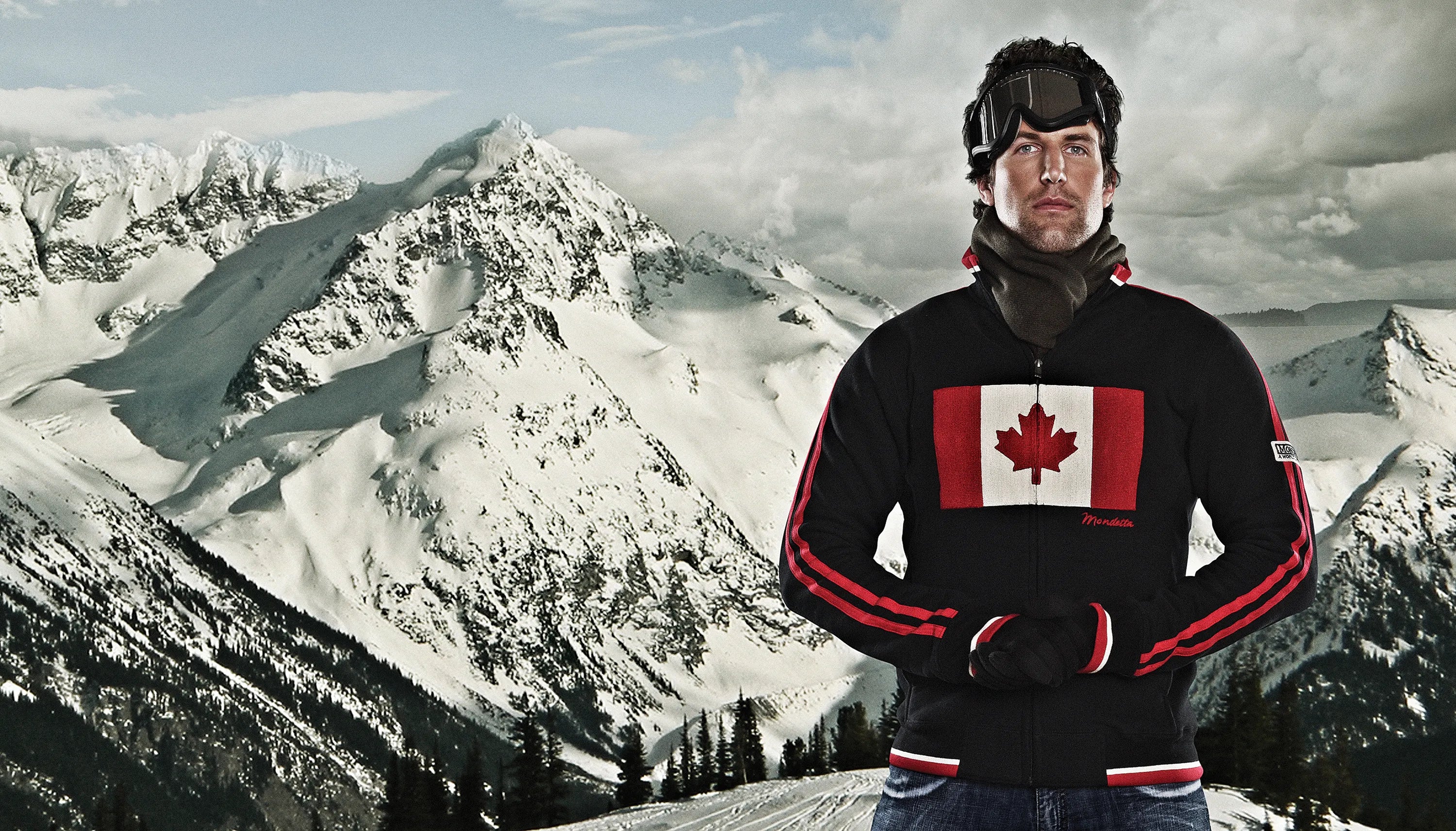 White man wearing black Mondetta Canada flag and red stripe zip-up sweater in front of mountain backdrop.