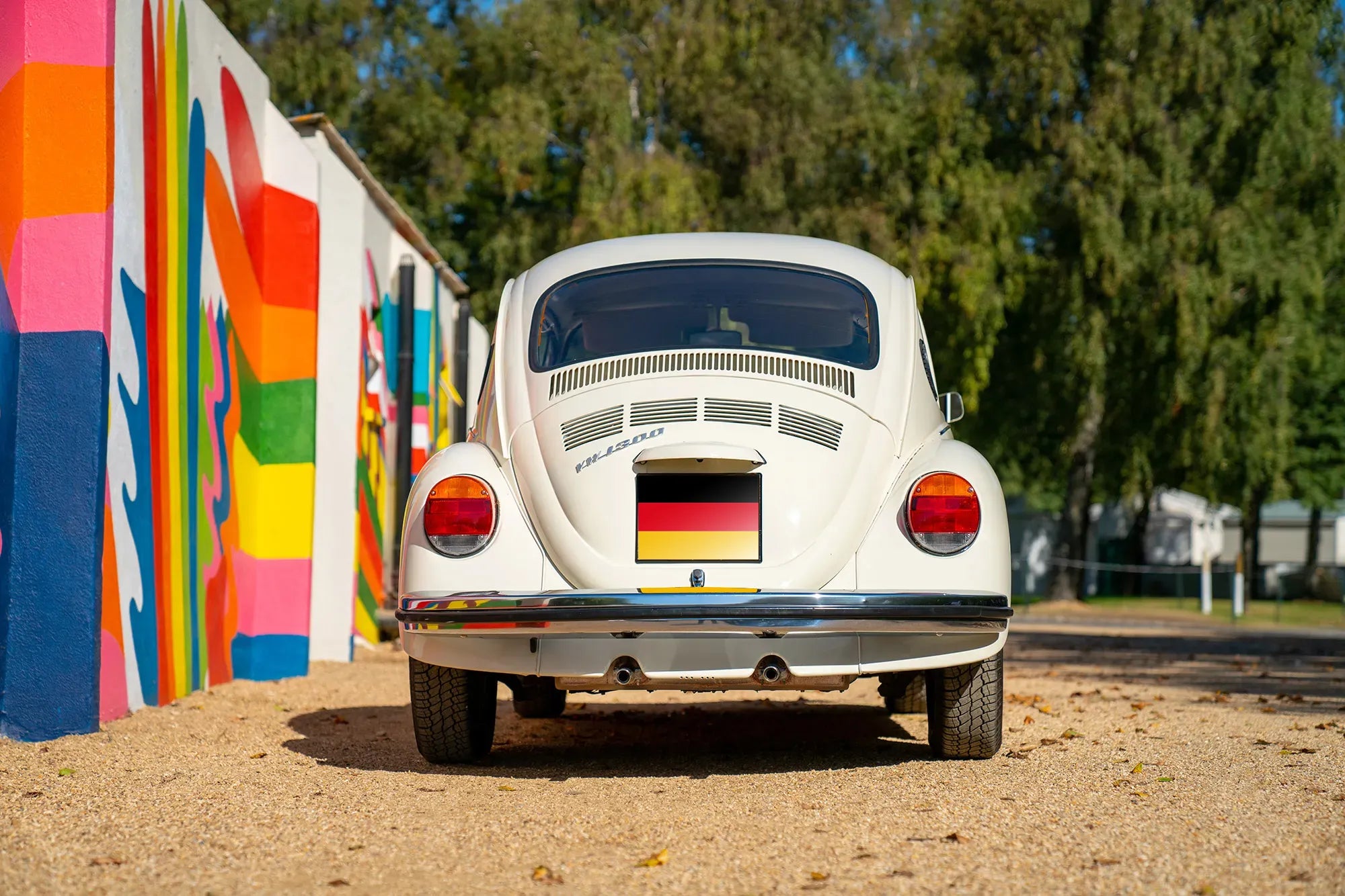 Vintage Volkswagen Beatle car with a German flag on the front parked next to trees and bright murals.